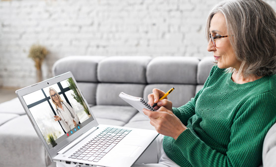 Older woman writing in notebook while video conferencing with doctor